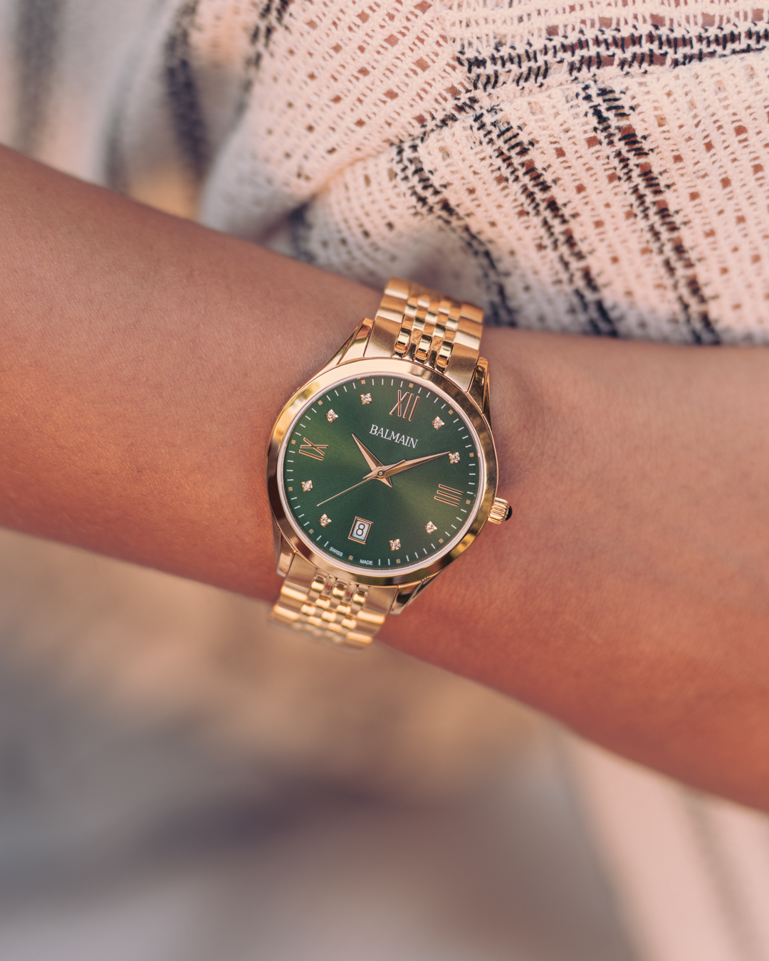 Close-up of a woman’s wrist in sunlight, wearing the Balmain Classic R B4310.31.75 watch. It has a 34mm round yellow gold PVD case, a green dial with 8 Top Wesselton diamonds, and a polished metal bracelet.