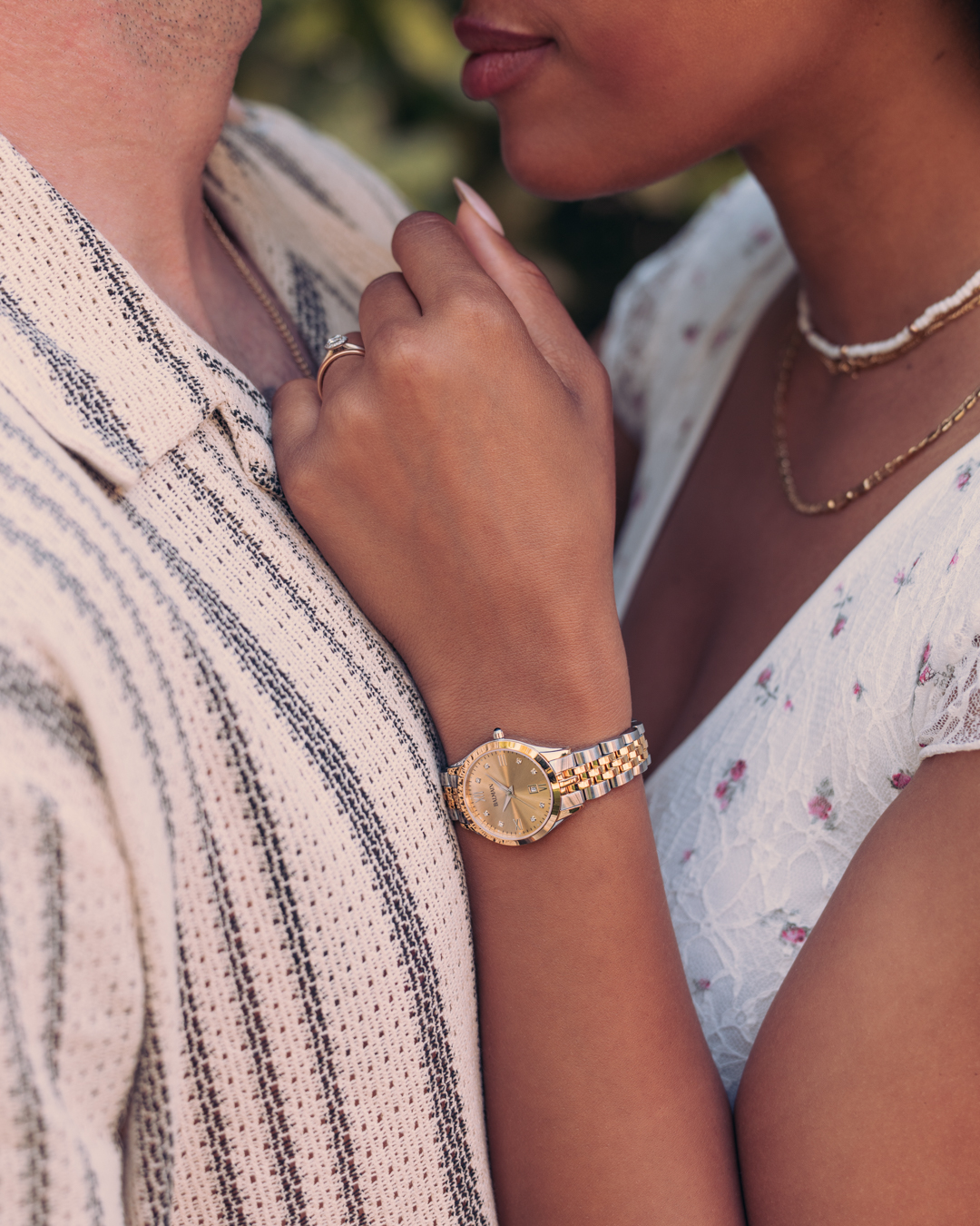 A stylish couple enjoys a sunny outdoor scene. The woman wears the Balmain Classic R B4112.31.25 watch. It has a 30mm round case made of stainless steel and yellow gold PVD. The watch features a yellow dial set with 8 Top Wesselton diamonds, along with a 